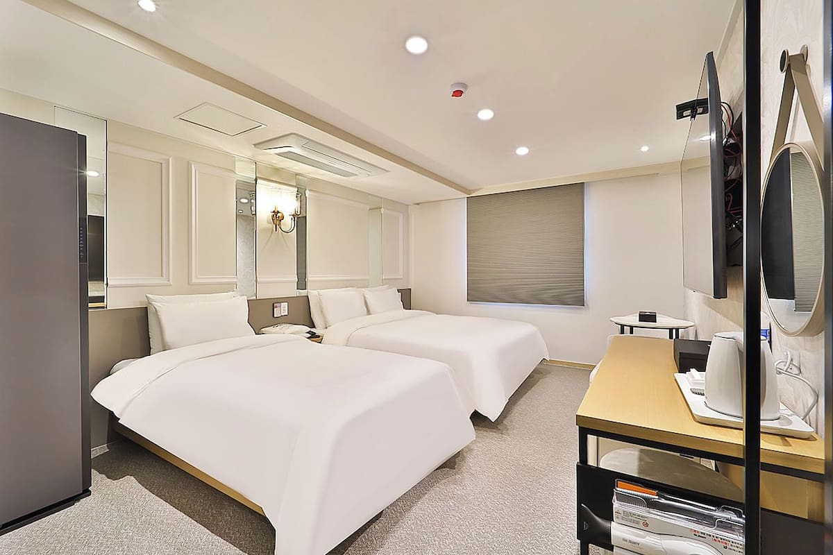 Ein Hotelzimmer mit zwei Betten, einem Schreibtisch mit Computer, einem Spiegel und einem Fenster mit Jalousien.
