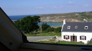 Vista costera desde el interior de un edificio, con una casa blanca de techo oscuro, cielo despejado y el mar al fondo.