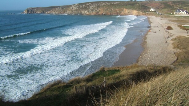 Un paisaje costero con playa, olas, zonas de hierba y edificios al fondo.