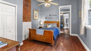 A bedroom with a wooden bed, a ceiling fan, a brick wall, and a view into another room.