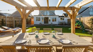 Eine hölzerne Pergola mit einem Esstisch, der für sechs Personen gedeckt ist, mit Blick auf ein Haus und einen Garten.