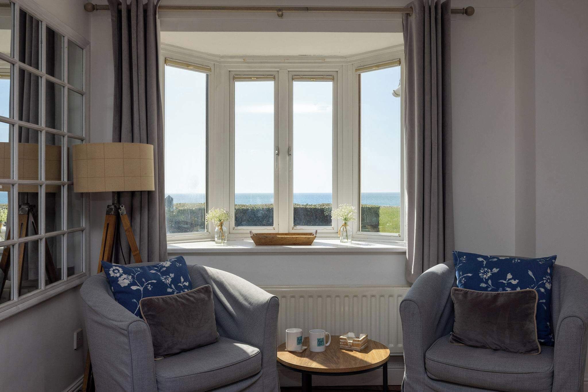 A room with two grey armchairs, a small wooden table, and a bay window with a view of the sea.