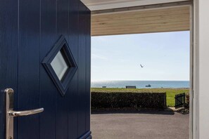 A view from a dark blue door to a beach with a bench, a hedge, and a boat in the distance. - Wonderful Seafront Home - Panoramic Ocean Views (Selsey)