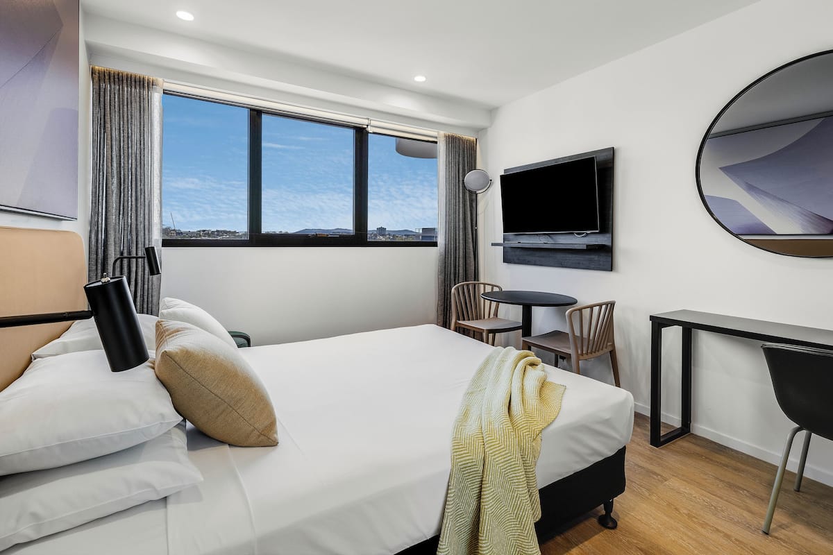 A hotel room with a bed, a desk, a chair, a TV, and a view of the city.