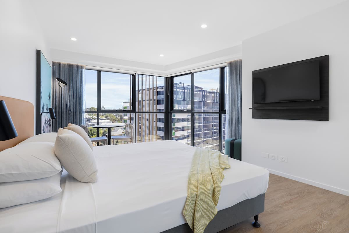 A modern hotel room with a large bed, a TV mounted on the wall, and a balcony with a view of the city.