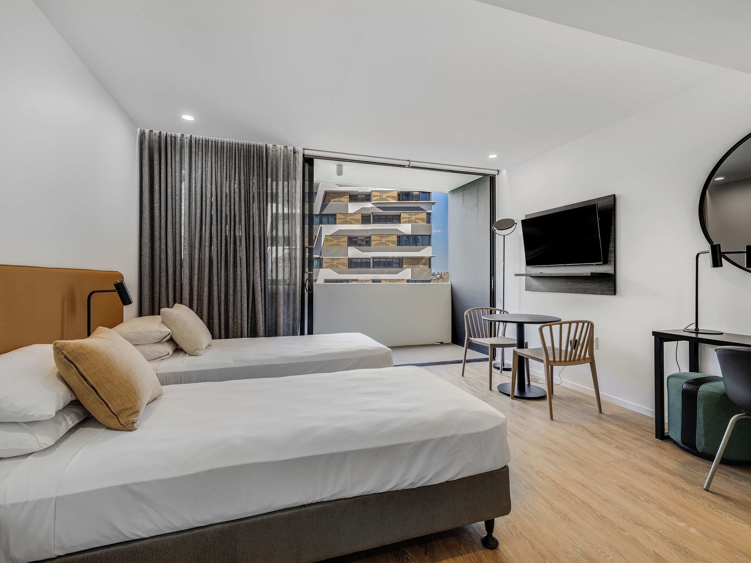 Een moderne hotelkamer met twee bedden, een bureau, een stoel, een televisie en een groot raam met uitzicht op een gebouw.