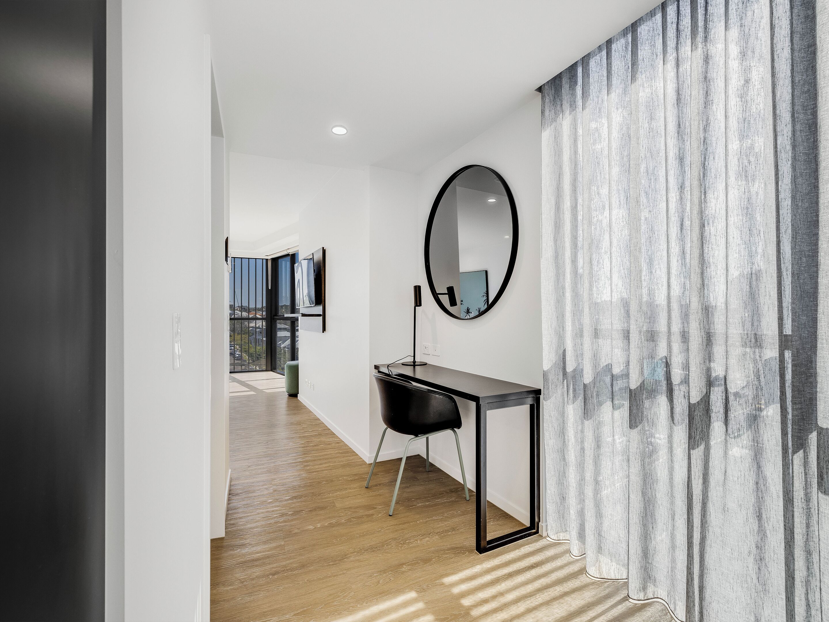 A modern hallway with a wooden floor, a black desk, a round mirror, and a view of the city through large windows.