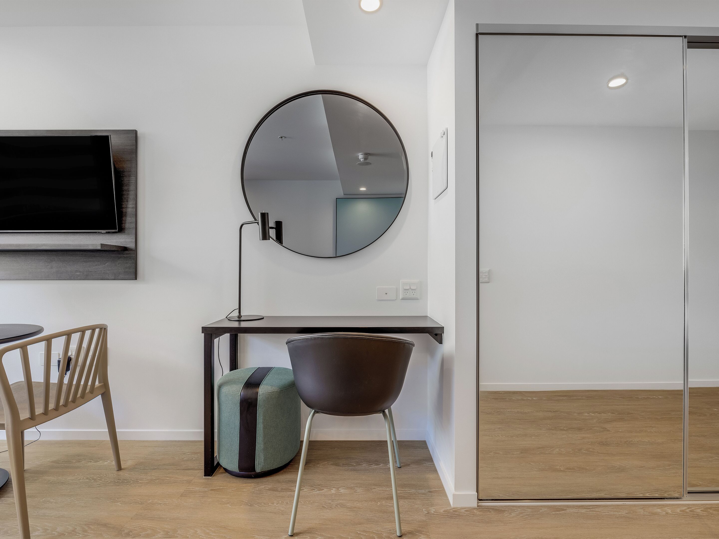 Une chambre moderne équipée d’un téléviseur à écran plat, d’un miroir rond, d’un bureau, d’une chaise et d’une armoire.