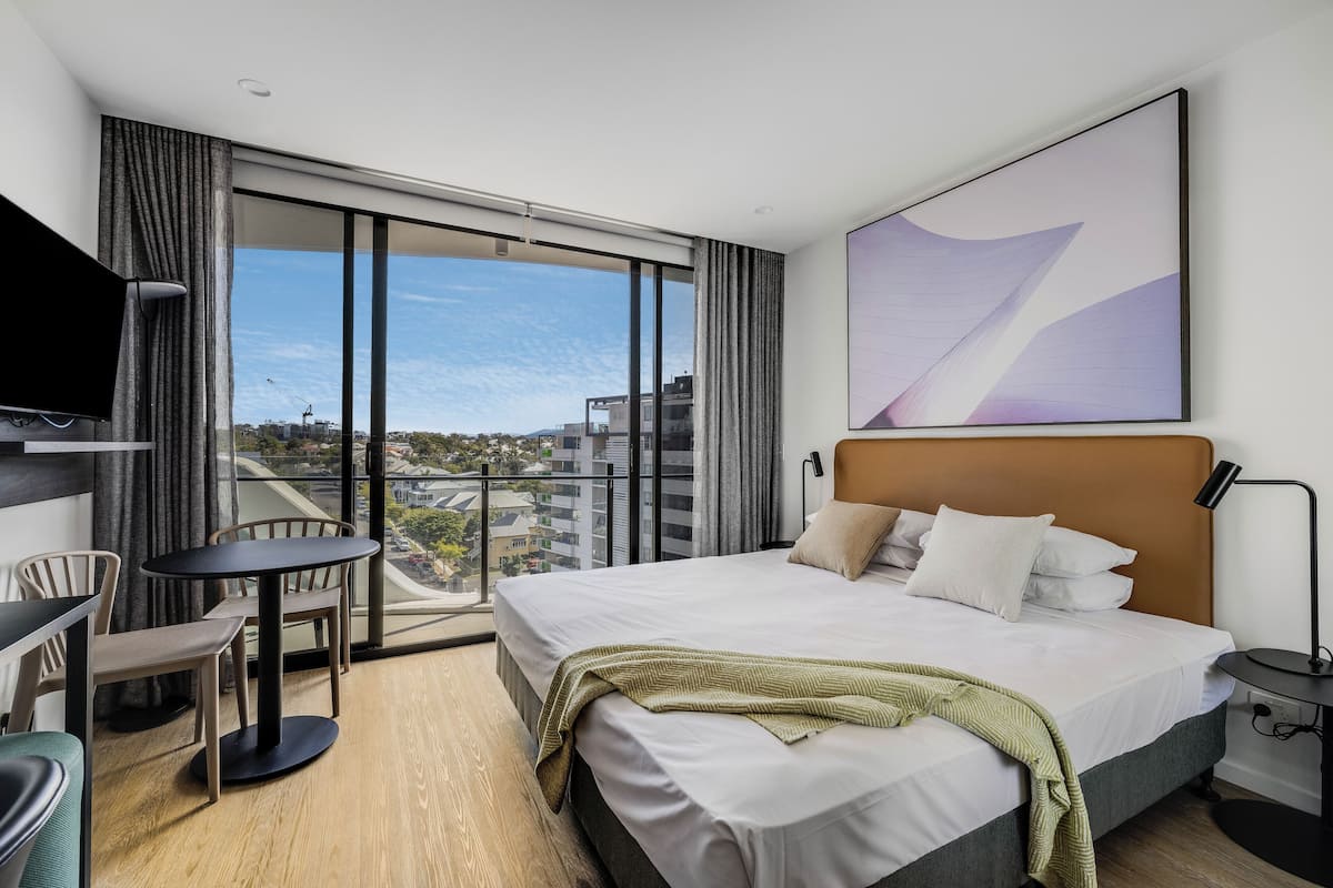 A modern hotel room with a large bed, a flat-screen TV, a dining area, and a balcony with a city view.