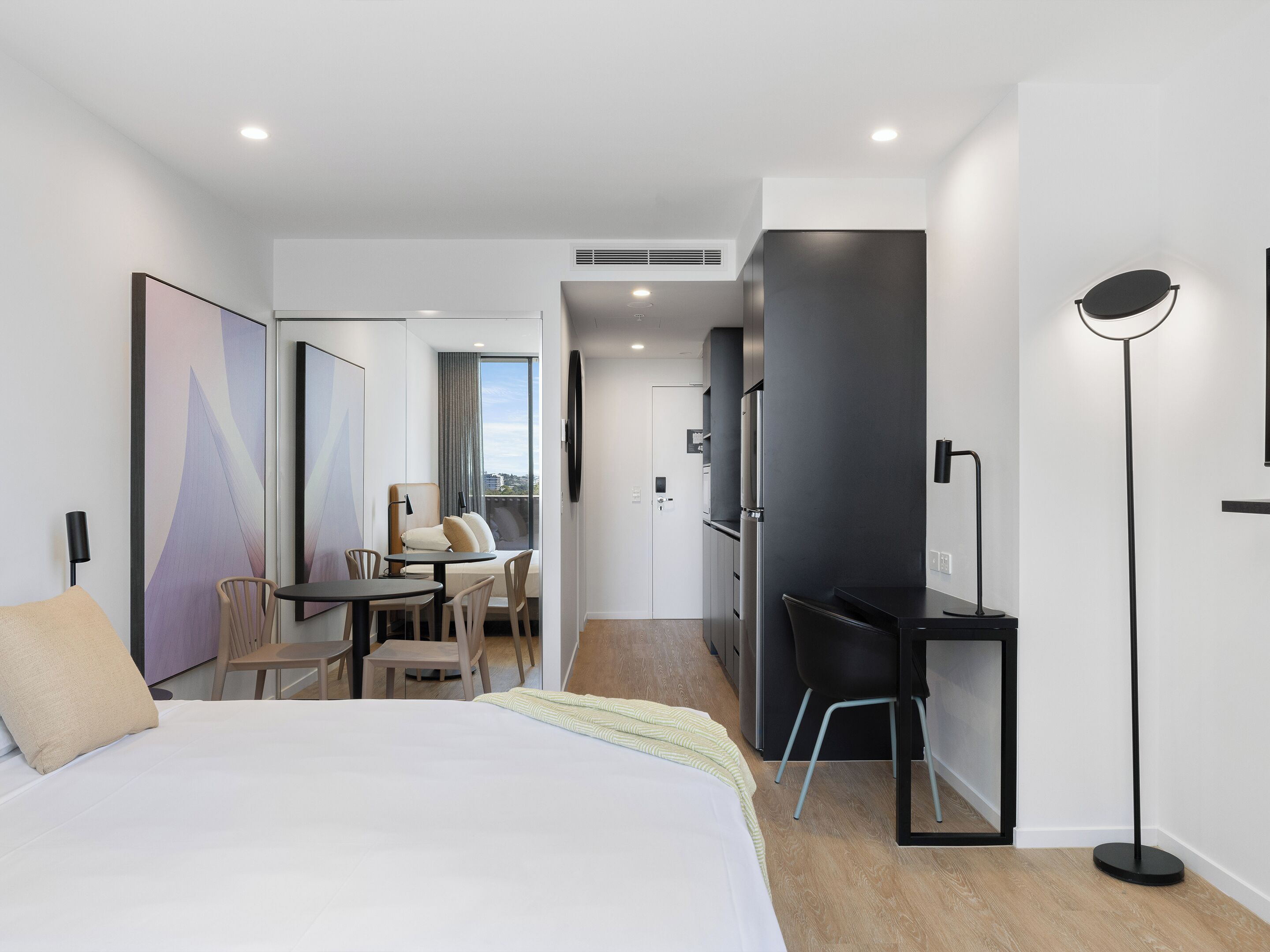 A modern hotel room with a large bed, a desk, a chair, a dining table, and a balcony with a view.