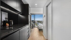 Una cocina moderna con armarios oscuros, una cafetera y un vaso de jugo en la encimera. Se ve un comedor con mesa y sillas, que conduce a un dormitorio con cama y vistas de la ciudad.