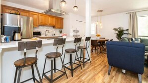 A modern kitchen with a central island, bar stools, and a dining area with a blue sofa. - Cozy Single-Story near Pineville Hospital, games (Charlotte)