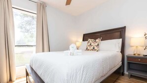 A neatly made bed with a white comforter and pillows, a bedside table with a lamp, and a window with curtains. - Cozy Single-Story near Pineville Hospital, games (Charlotte)