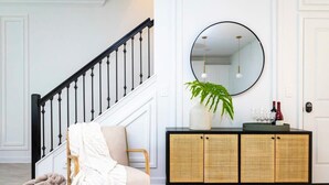 Un escalier avec des balustrades noires, une chaise en osier recouverte d’une couverture blanche, une console en osier avec un miroir et une plante.