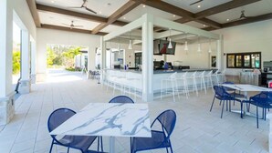 Un espace de restauration en plein air avec des tables blanches et des chaises bleues, un bar avec des tabourets blancs et des ventilateurs de plafond.