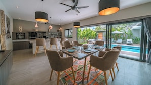 Una cocina moderna con armarios oscuros, un comedor con una mesa grande y sillas, y vista a una piscina a través de amplios ventanales.
