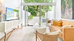 A modern living room with a large TV, comfortable seating, and a view of lush greenery through glass doors.
