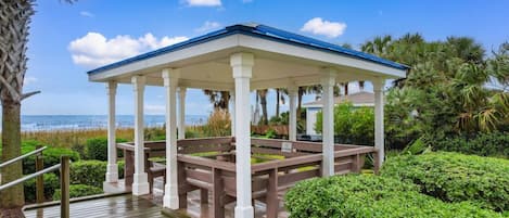 A covered outdoor seating area with a wooden deck, white columns, and a view of the beach.