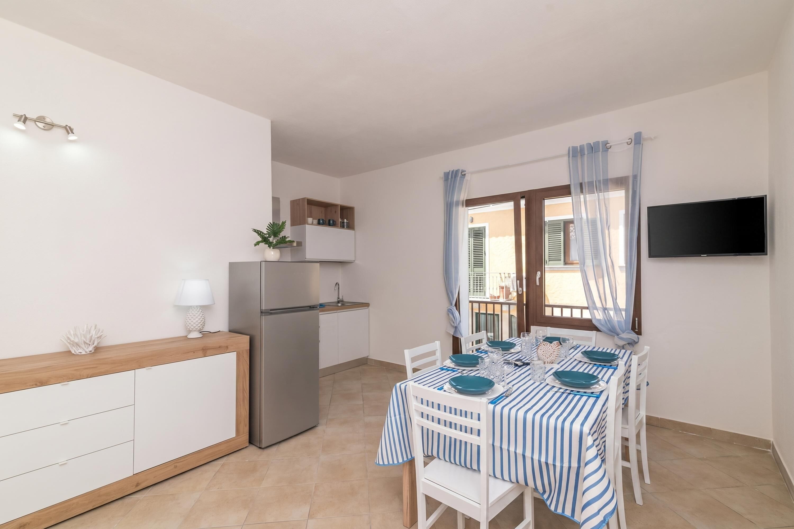 A compact kitchen with a refrigerator, a dining table set for four, and a TV mounted on the wall.