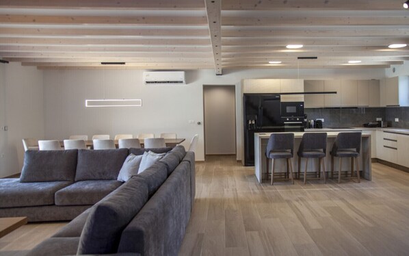 Una sala de estar moderna con un sofá seccional gris, un comedor con mesa y sillas, y una cocina con refrigerador negro, microondas y taburetes de bar.