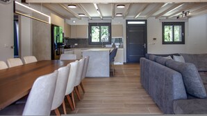 A modern kitchen with a central island, a dining area with a wooden table and chairs, and a grey sofa in a living area. - Villa Olia in Agios Petros (Lefkada)