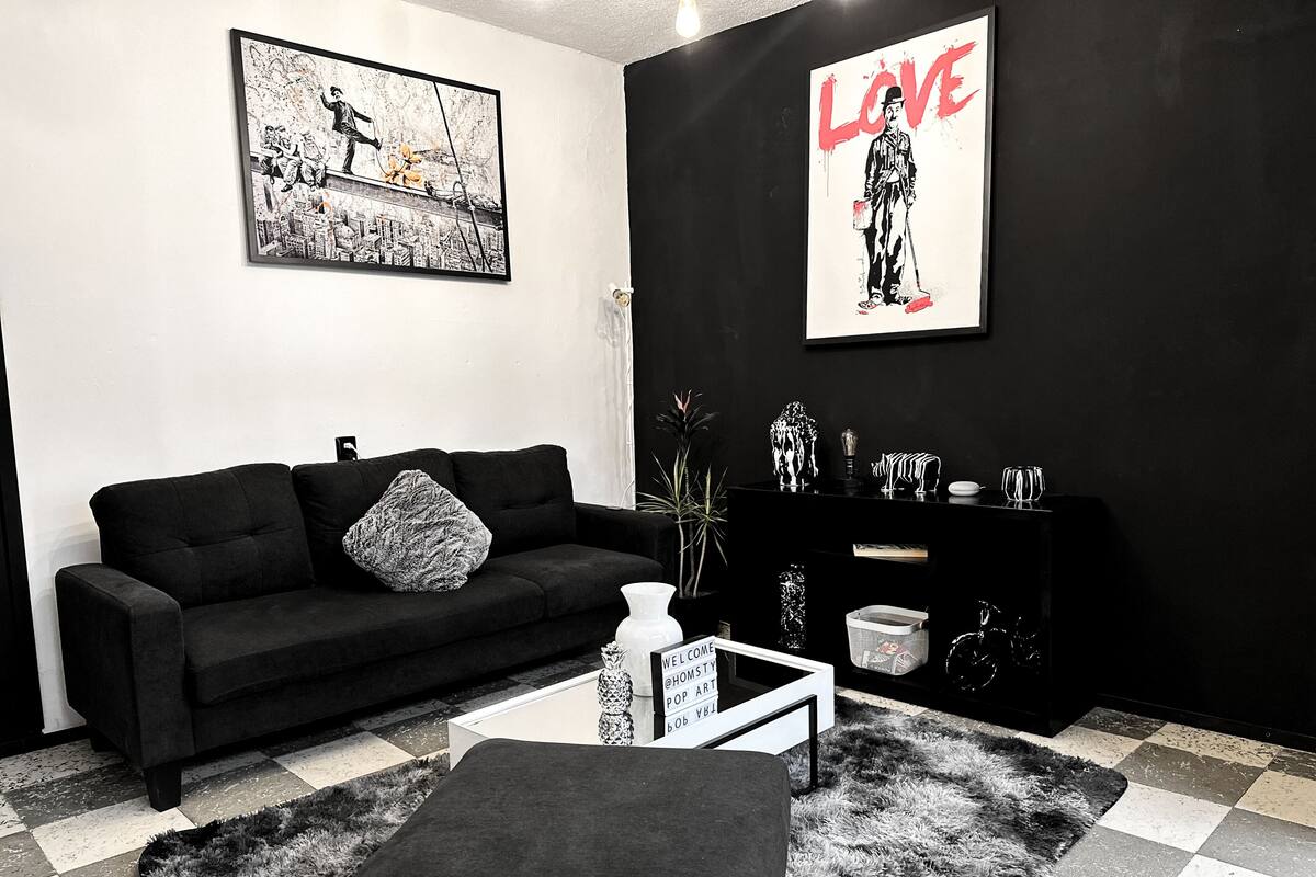 A modern living room with a black sofa, a white coffee table, and a black and white rug. There are two framed artworks on the walls and a black cabinet with decorative items.