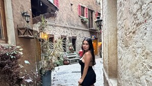 Un estrecho callejón con pavimento adoquinado, macetas con plantas y edificios tradicionales con contraventanas rojas.