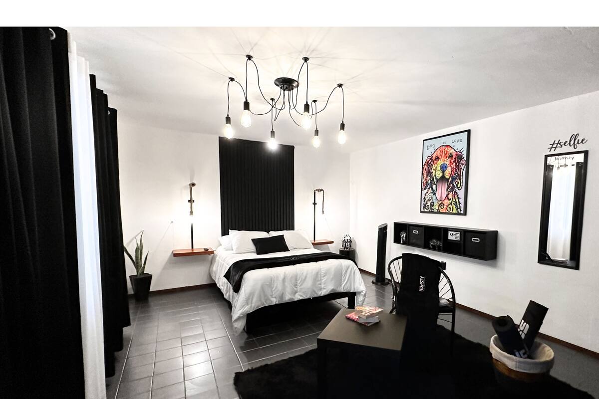A modern bedroom with a bed, bedside tables, a desk, and a chair. There is a chandelier, a painting, and a mirror on the wall.