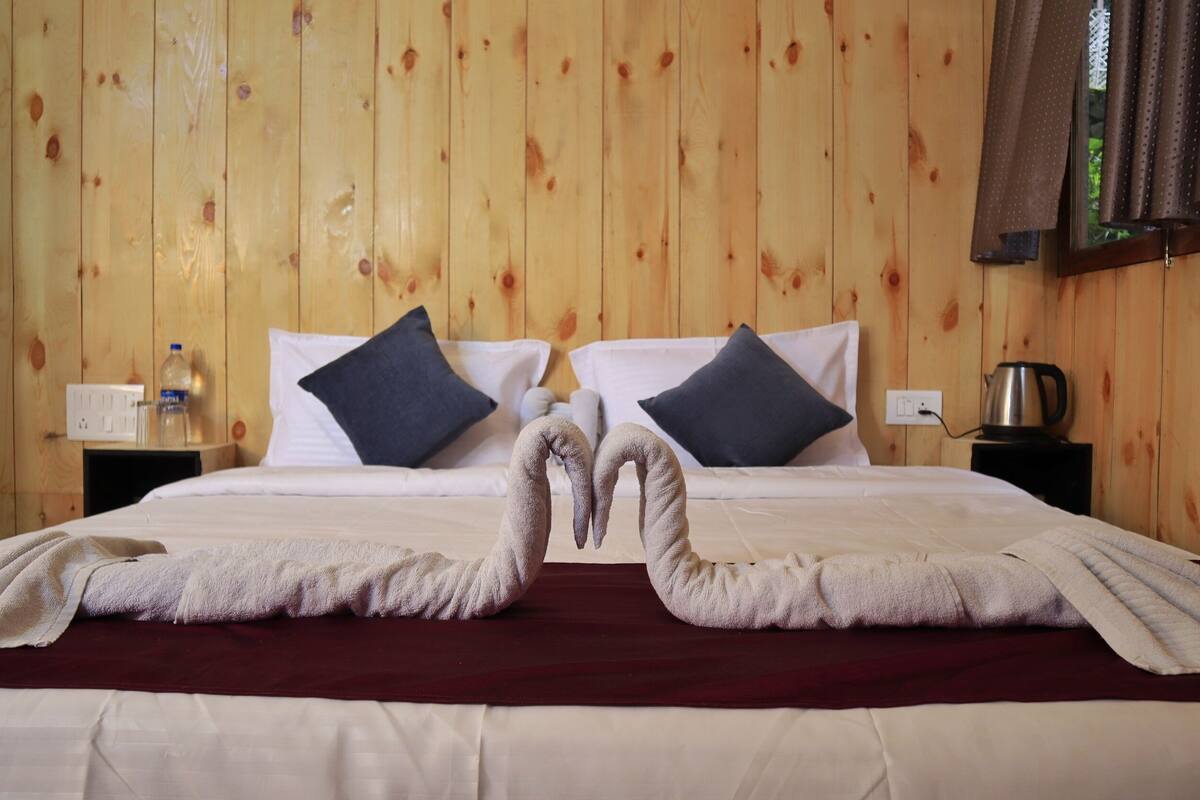 Ein Hotelzimmer mit einer Wand aus Holzpaneelen, einem Bett mit weißen Leinentüchern, zwei dunklen Kissen und einer roten Bettdecke. Auf dem Bett liegen zwei Handtücher in Herzform.