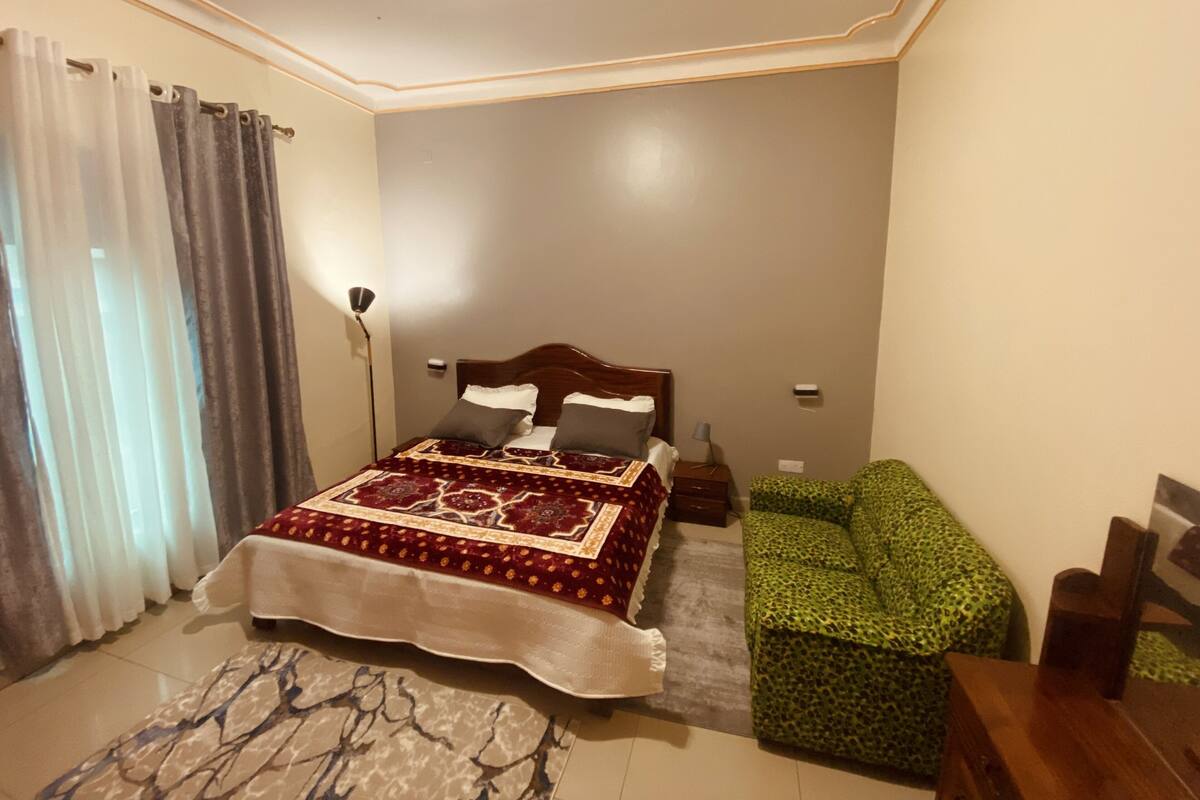 A bedroom with a bed, a green patterned sofa, a floor lamp, and a patterned rug.