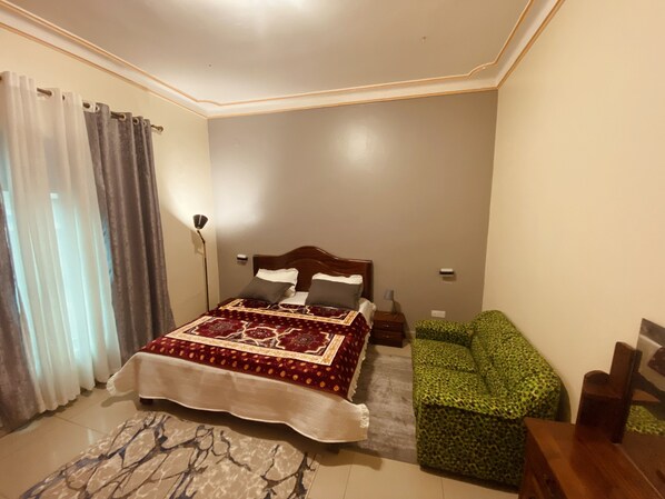 A bedroom with a bed, a green patterned sofa, a floor lamp, and a patterned rug. - Affordable Homes Kigali (Kigali)