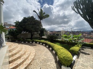 A landscaped area with a curved pathway, a palm tree, and a view of a residential area in the background. - Affordable Homes Kigali (Kigali)