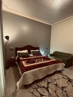 A bedroom with a bed, bedside table, lamp, and a green patterned chair. - Affordable Homes Kigali (Kigali)