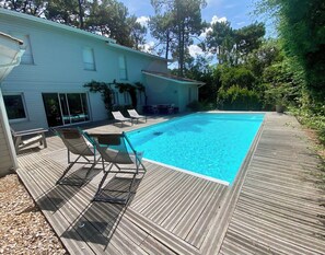 Een zwembad met een houten terras, ligbedden en een huis op de achtergrond.