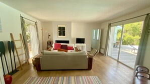 Ein großzügiger Wohnraum mit einer großen Couch, einem Fernseher und Blick ins Freie durch große Fenster.