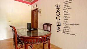 A dining area with a round glass table and wooden chairs, a door in the background, and a wall with a 'WELCOME' sign listing various languages. - Hotel O Kedai Cinta Guest House (West Cirebon)