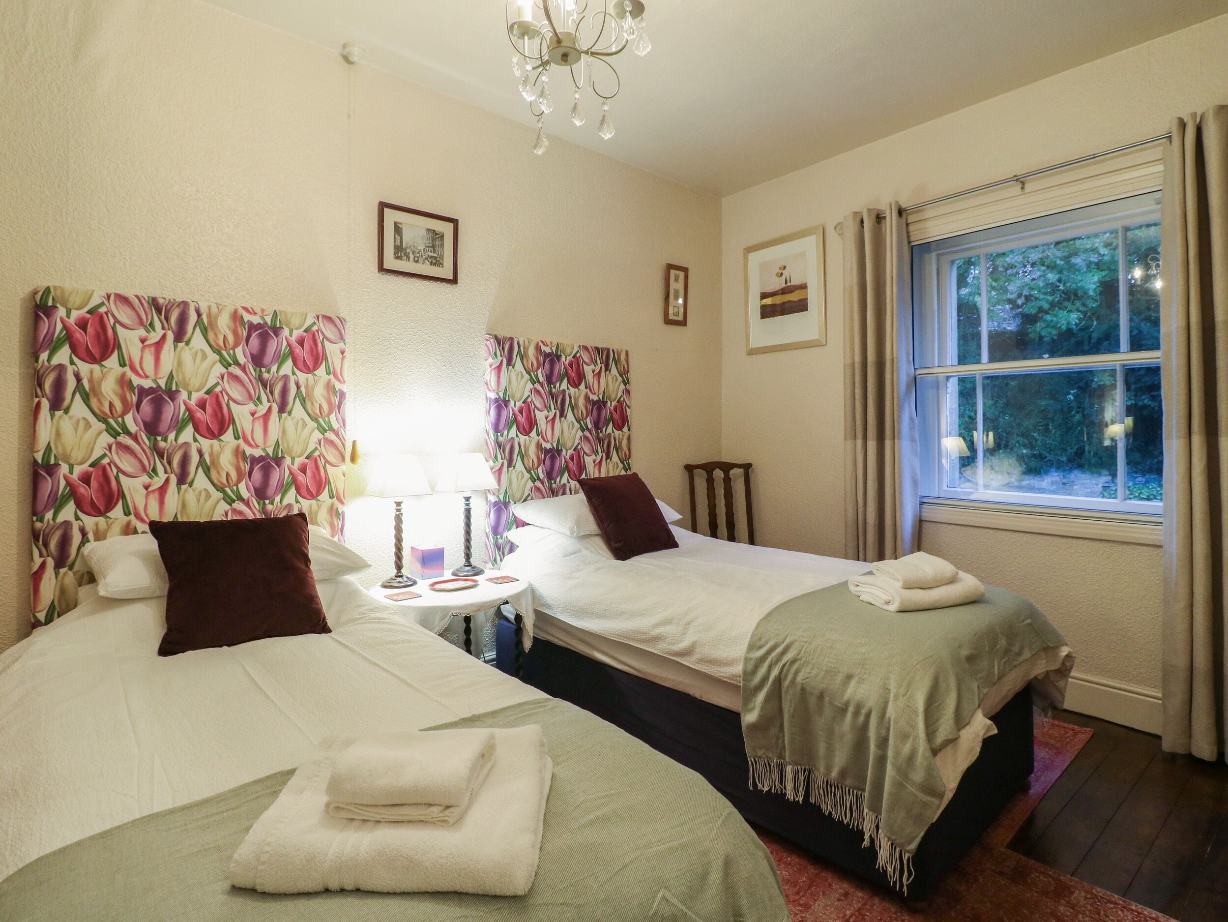 A hotel room with two beds, floral headboards, a small round table with lamps, and a window with curtains.