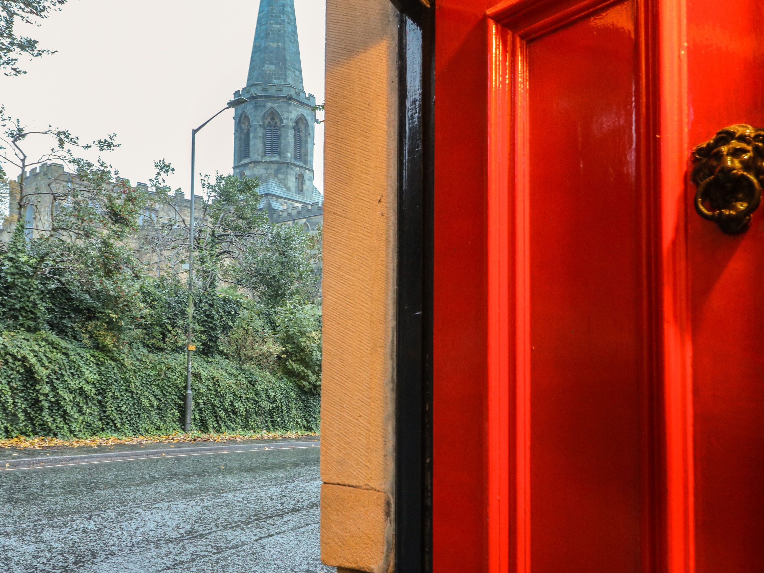 A red door with a brass knocker, a street view with a church spire in the background, and a hedge along the sidewalk.