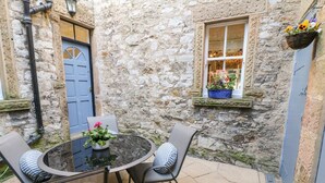 A stone building with a round table and chairs, a blue door, and a window with a flower box. - Knoll Cottage (Bakewell)