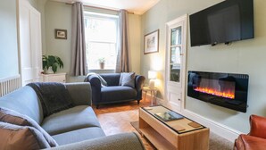 A living room with a sofa, a coffee table, a fireplace, a TV, and a window with curtains. - Knoll Cottage (Bakewell)