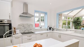 Een moderne keuken met witte kasten, een roestvrijstalen afzuigkap, een inbouwoven, een raam met blauwe decoratielijsten en een groot raam met uitzicht op groen.