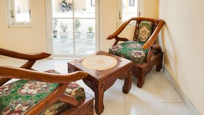 A room with two wooden chairs with floral upholstery, a small wooden table with a decorative center, and a view of the street through a glass door. - OYO Life  91569 Omah Wijaya Kusuma Syariah (Asrikaton)