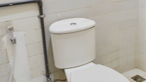 A white toilet with a tank and a bidet attachment in a bathroom with white tiled walls and floor. - OYO Life  91569 Omah Wijaya Kusuma Syariah (Asrikaton)
