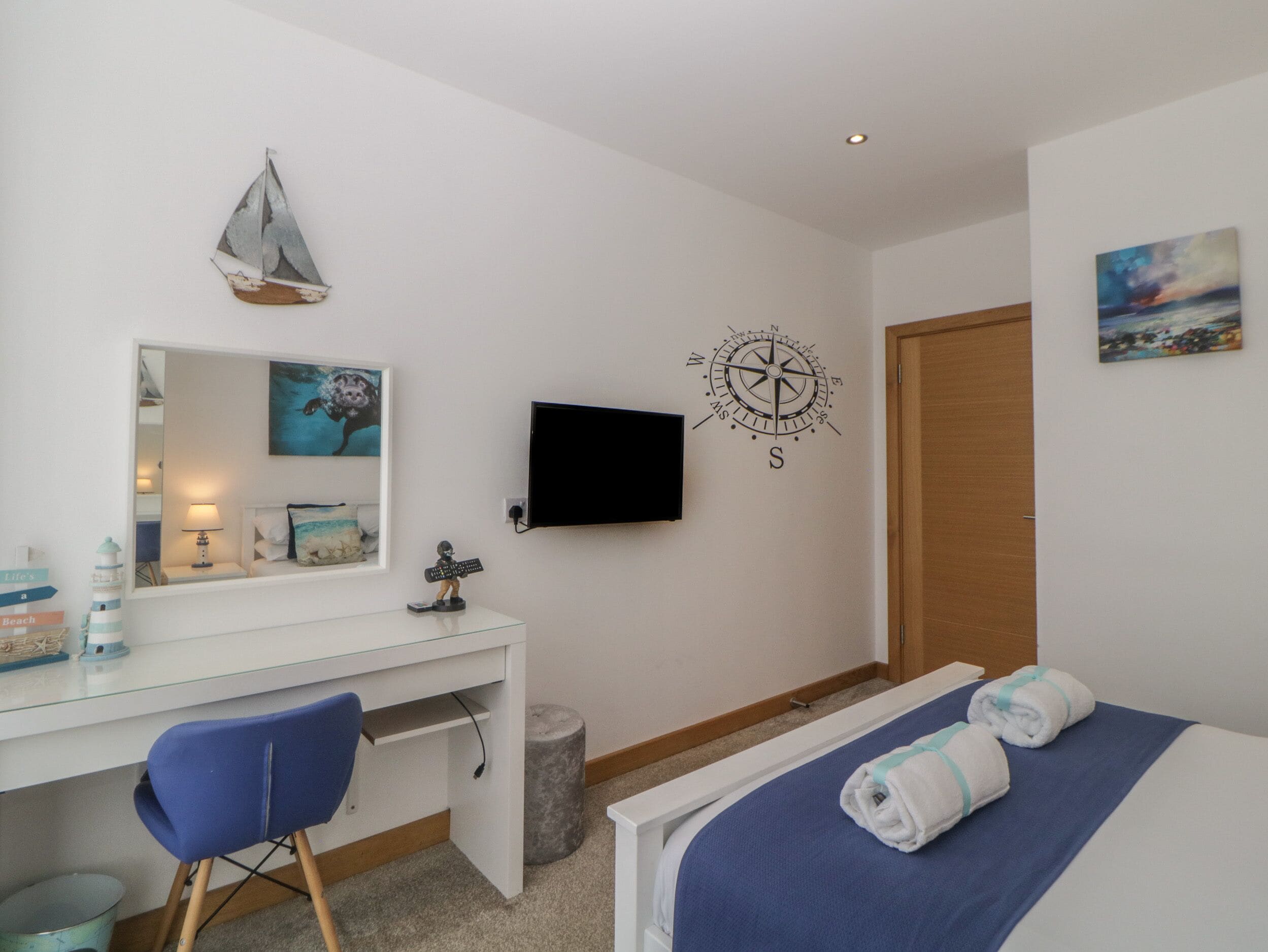 A bedroom with a bed, a desk with a chair, a TV, a compass wall decoration, and a door.