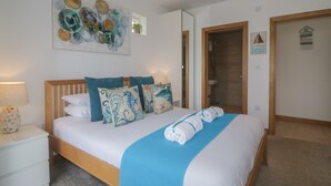 Una cama bien hecha con una manta azul y almohadones decorativos, una mesita de noche con una lámpara y una obra de arte en la pared.