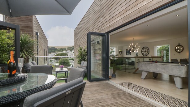 Un espacio exterior moderno para comer con mesa y sillas de vidrio, una botella de champán y vistas a la piscina y la zona de la piscina.
