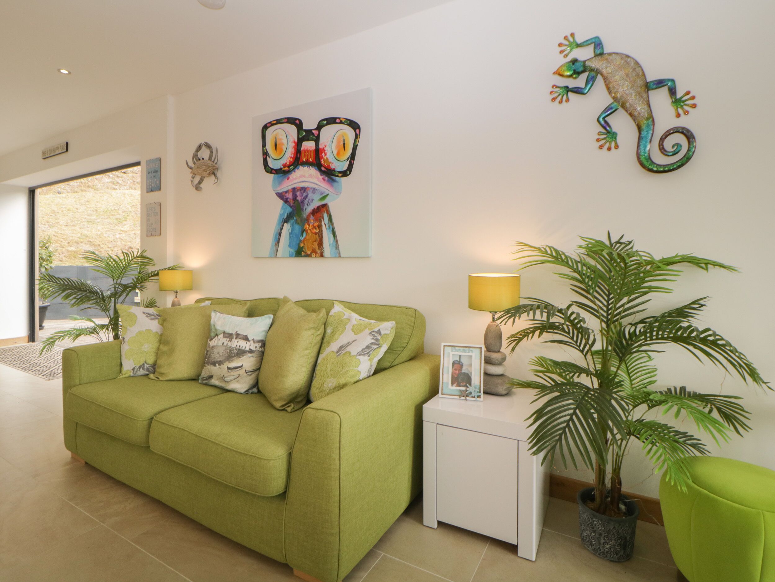 A green sofa with decorative pillows, a small white nightstand with a lamp, and a framed photo. There are two potted plants and a colorful artwork on the wall.