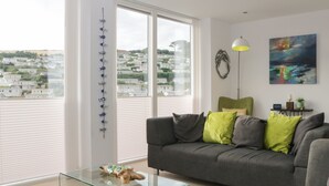Un salón moderno con un sofá gris, cojines verde y gris, una mesa de centro de vidrio y vistas a una zona residencial a través de grandes ventanales.