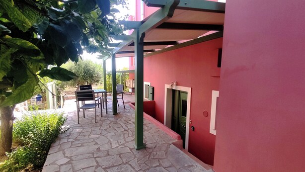 A stone-paved pathway leading to a building with a red exterior. There are two tables and chairs under a green pergola. - Quiet apartment in Marathonas - 5 min from the beach! (Μαραθώνας)