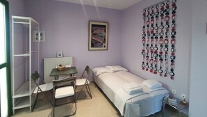 A small, well-lit room with a bed, a desk, and a chair. There is a radiator, a window, and a wall-mounted light fixture. - Quiet apartment in Marathonas - 5 min from the beach! (Μαραθώνας)